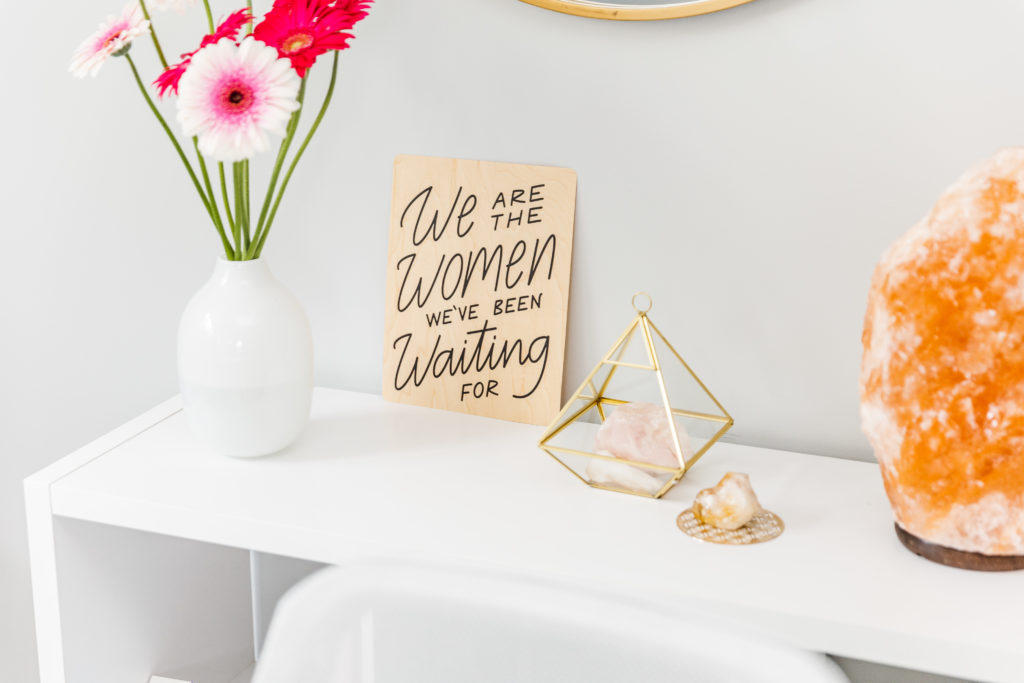 A bookshelf with a women's sign, orange salt lamp, geometric shape, and flowers. 