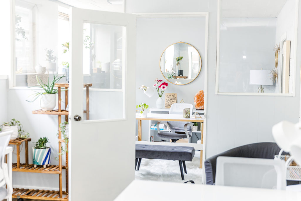 An open door office space with a mirror, desk, bench, plant stands and neutral paint colors. 